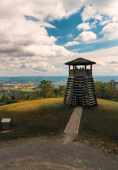 A wooden observation tower on a grassy hill, surrounded by trees and mountains under a cloudy sky.