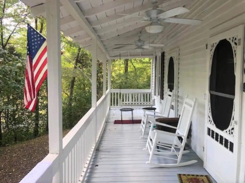 A porch with rocking chairs, a small table, and an American flag, surrounded by greenery.