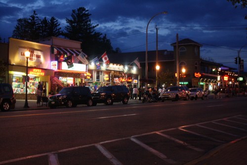 A street scene at dusk with shops, flags, and parked cars, illuminated by streetlights and colorful storefronts.