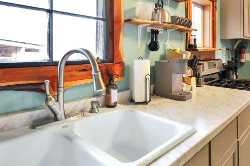 A modern kitchen sink with a silver faucet, marble countertop, and wooden shelves holding kitchen items and a coffee maker.