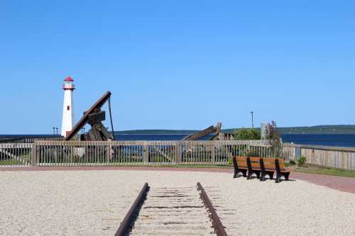 A lighthouse stands by the water, with benches and old railway tracks in the foreground against a clear blue sky.