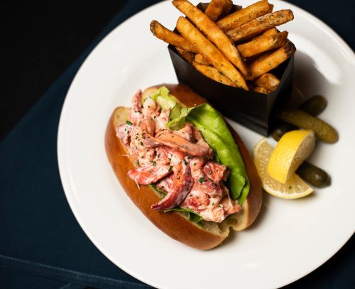 A lobster roll with lettuce on a bun, served with fries, pickles, and a lemon wedge on a white plate.