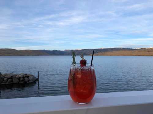 A refreshing cocktail garnished with rosemary and a cherry, set against a serene lake and mountain backdrop.