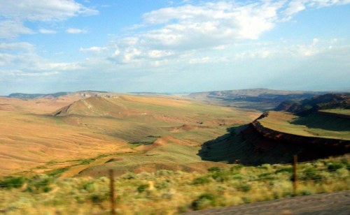 A vast, arid landscape with rolling hills and valleys under a blue sky with scattered clouds.