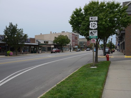 A quiet street scene featuring shops, a "West 12" road sign, and trees lining the sidewalk on a cloudy day.