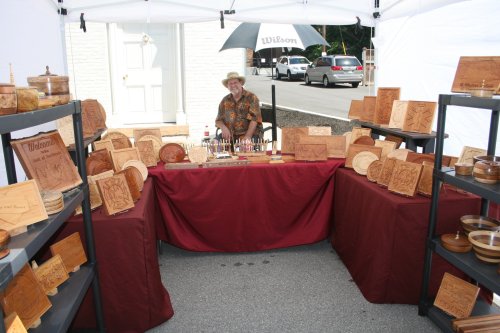 A vendor stands behind a table displaying various wooden crafts under a tent, with a "Welcome" sign above.
