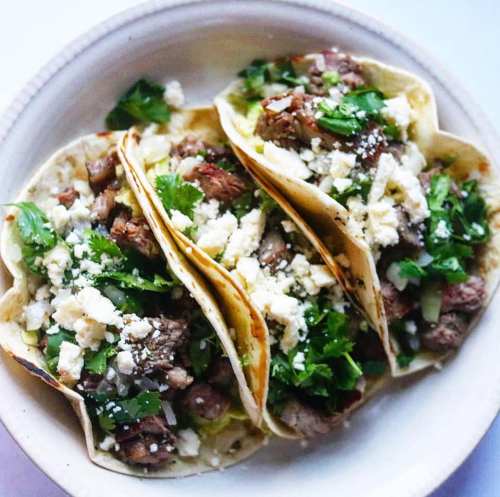 Three tacos filled with seasoned meat, topped with cilantro and crumbled cheese, served on a white plate.