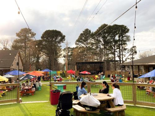A lively outdoor space with people dining at picnic tables, colorful umbrellas, and green grass under a cloudy sky.