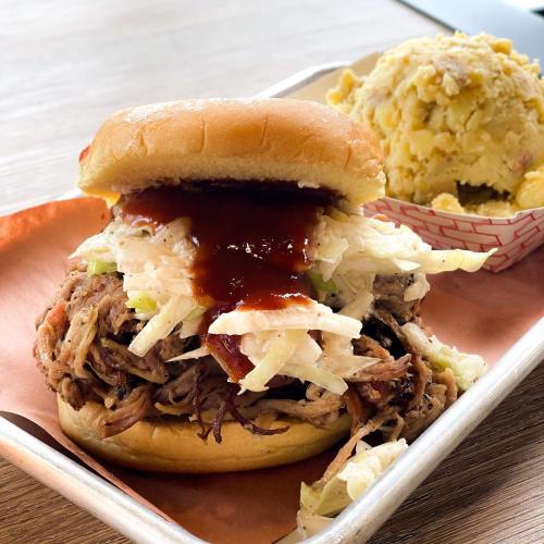A pulled pork sandwich topped with coleslaw and barbecue sauce, served with a side of potato salad.