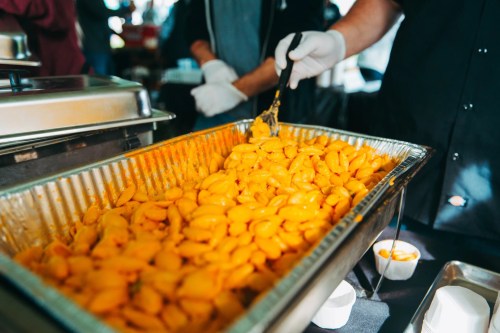 A tray filled with bright yellow macaroni and cheese, being served by a person wearing gloves.