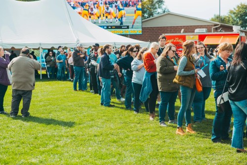 A long line of people waiting outside near a large screen and food trucks on a sunny day.