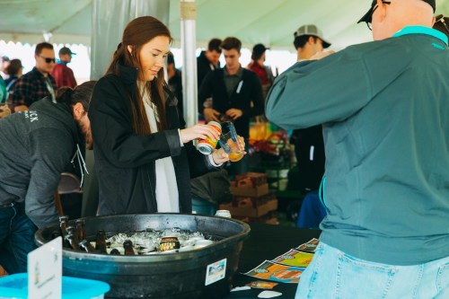 A woman pours a drink from a can into a cup at a crowded outdoor event under a tent.