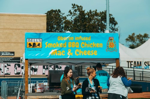A food stall with a blue banner advertising Oberon infused smoked BBQ chicken mac and cheese at an outdoor event.