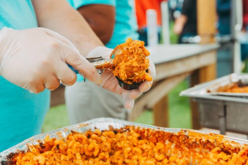 A person in gloves serves a portion of macaroni and cheese from a large tray at an outdoor event.