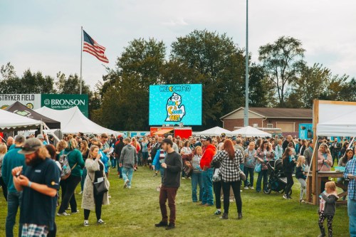 A large crowd gathers at an outdoor event with tents, a big screen displaying "Welcome to Fest," and an American flag.