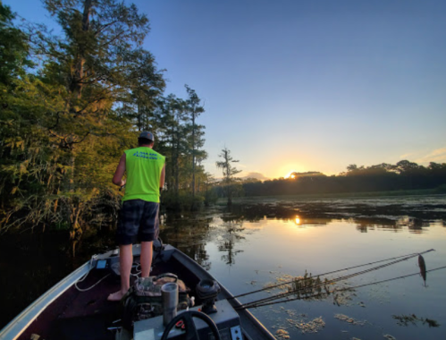 A person fishing from a boat at sunrise, surrounded by trees and calm water reflecting the colorful sky.