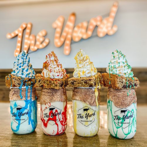 Four colorful milkshakes topped with whipped cream and drizzled with sauces, displayed in jars with "The Yard" sign in the background.