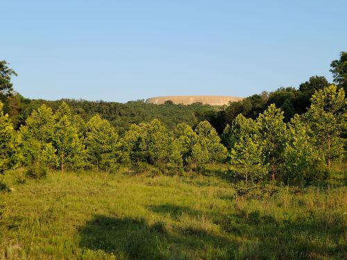 A view of a green landscape with trees, featuring a large, distant structure on a hill under a clear blue sky.