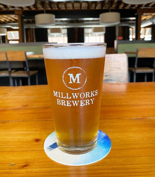 A glass of beer with the logo "Millworks Brewery" on a wooden table, with a blurred background of a brewery interior.