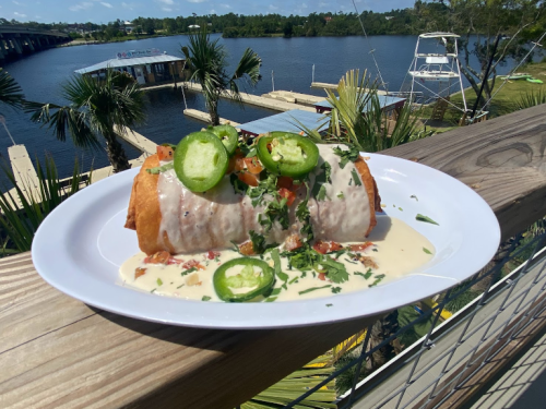 A plate of a large, crispy burrito topped with cheese sauce, jalapeños, and fresh herbs, with a scenic waterfront view.