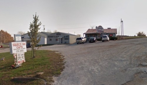 A gravel road leads to a small business area with a café and a barbershop, surrounded by trees and open space.