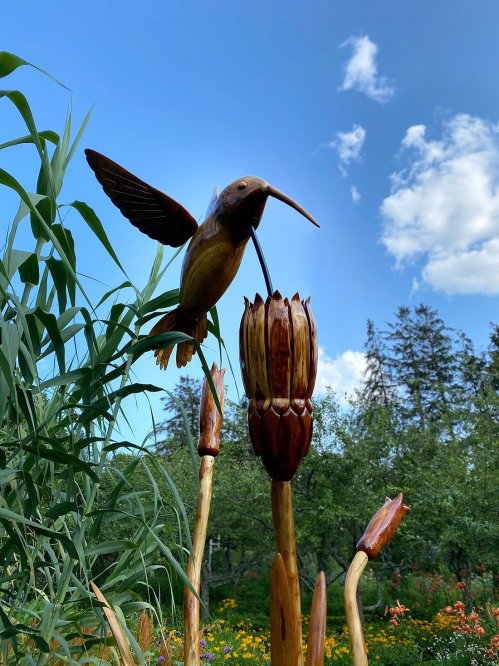 A wooden hummingbird sculpture hovering over a carved flower, set against a blue sky and green trees.