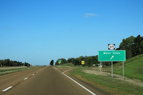 Road sign for Water Valley on a highway, with clear blue sky and green trees in the background.