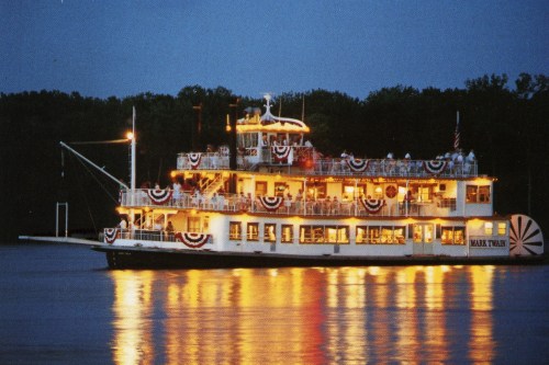 A lit riverboat named "Mark Twain" adorned with red, white, and blue decorations, reflecting on the water at dusk.