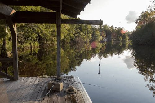 A serene river scene from a wooden dock, featuring fishing gear and lush greenery reflected in the calm water.