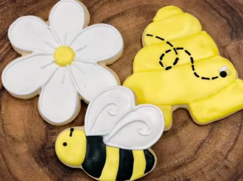 Three decorated cookies: a white flower, a yellow honeycomb, and a black-and-yellow bee, all on a wooden surface.