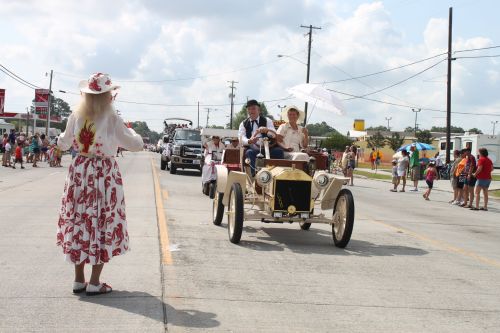 A vintage car parade with participants in period costumes, a woman in a hat waves, and spectators line the street.