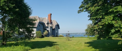 A large gray house with a chimney, surrounded by trees and a green lawn, overlooking a calm body of water.