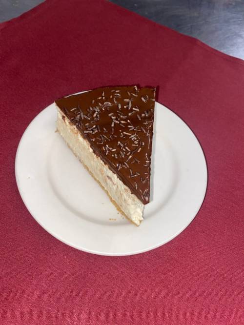 A slice of cheesecake topped with chocolate glaze and chocolate shavings, served on a white plate.