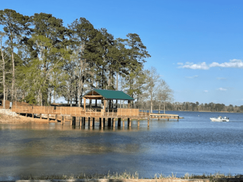 A wooden dock with a green-roofed gazebo by a calm lake, surrounded by trees under a clear blue sky.