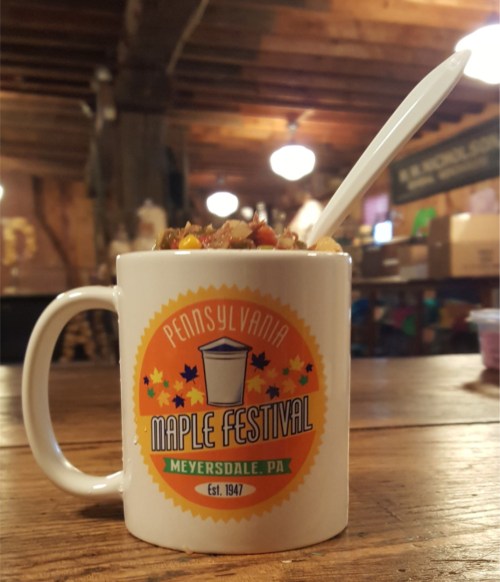 A white mug with a colorful "Pennsylvania Maple Festival" logo, filled with a dessert and a spoon, on a wooden table.