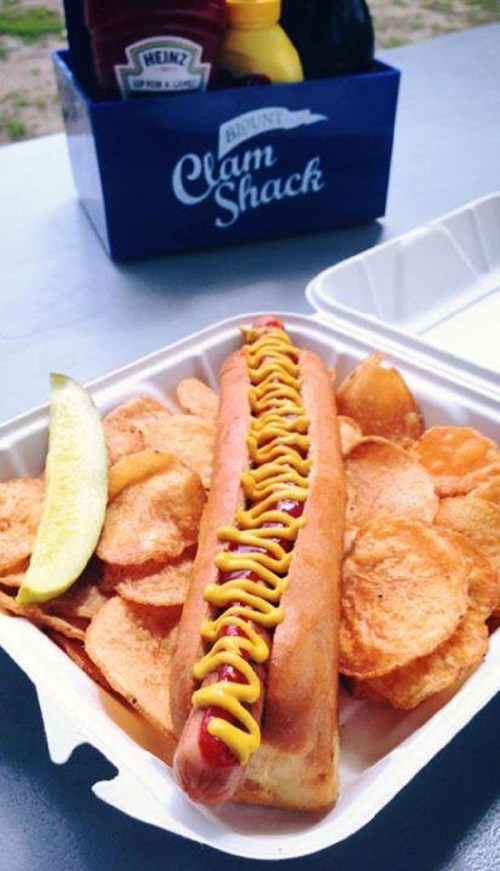 A hot dog with mustard and ketchup served with potato chips and a pickle slice in a takeout container.