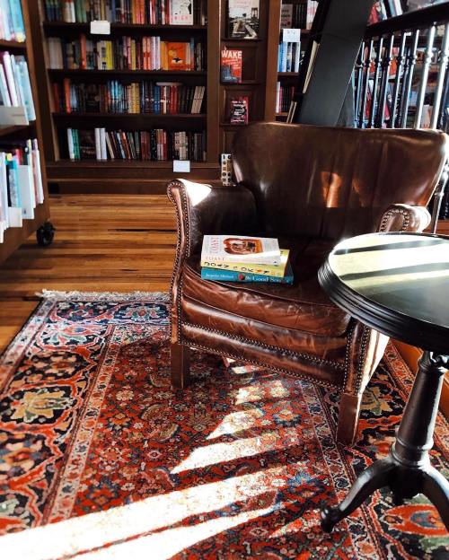 A cozy leather armchair beside a small table, with books stacked on the floor, in a warmly lit bookstore.