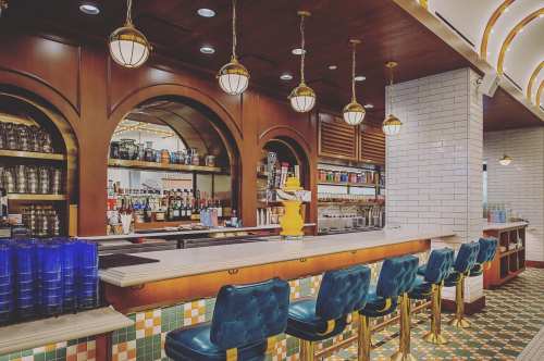 A stylish bar with blue stools, wooden accents, and decorative lighting, featuring shelves of drinks and a tiled counter.