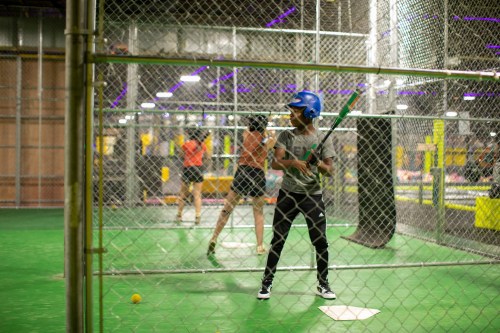 A young man in a blue helmet stands ready to bat in a batting cage, while another person swings in the background.
