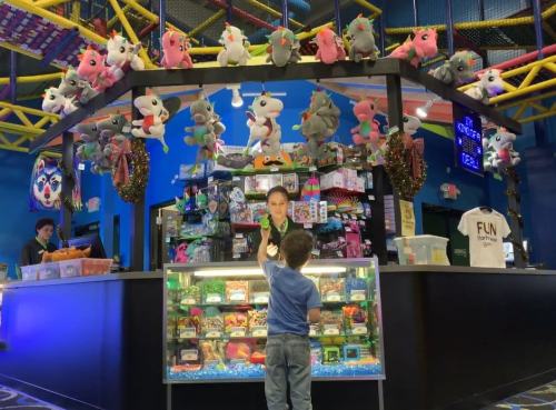 A child points at a prize counter filled with toys and stuffed animals, while an attendant stands behind the display.