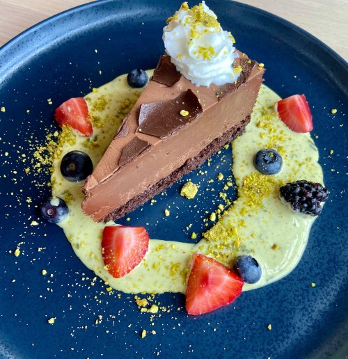 A slice of chocolate mousse cake topped with whipped cream, surrounded by berries and a creamy sauce on a blue plate.