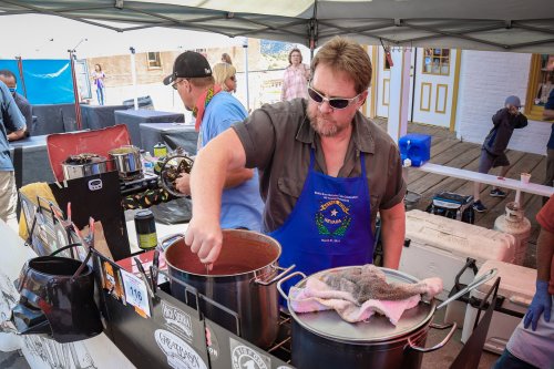 A man in sunglasses stirs a pot at a food stall, while another person works nearby at a festival.