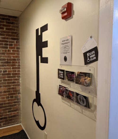 A large black key silhouette on a wall next to informational flyers and a fire alarm in a brick-walled room.