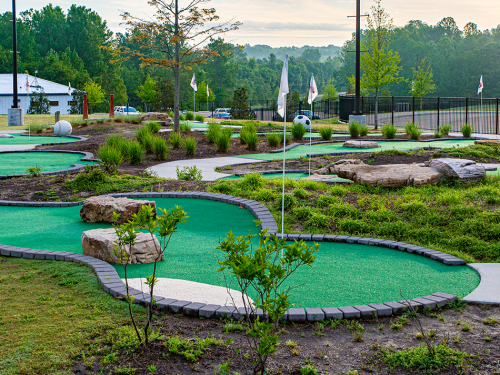 A scenic mini-golf course with green turf, rocks, and flags, surrounded by trees and a misty landscape in the background.