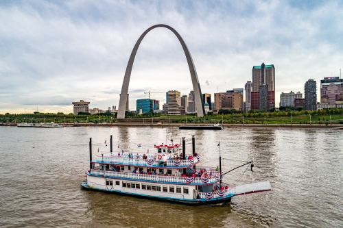 A decorated riverboat sails on the water with the St. Louis Arch and city skyline in the background.