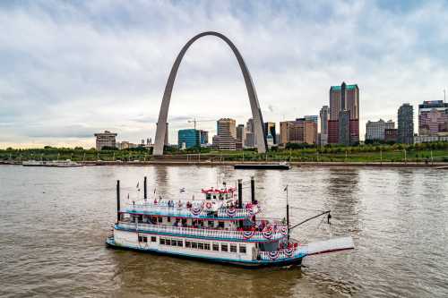 A decorated riverboat sails on the water with the St. Louis Arch and city skyline in the background.