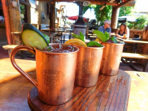 Three copper mugs filled with cocktails, garnished with cucumber, lemon, and mint, on a wooden table in a sunny outdoor setting.