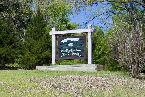 Sign for Woolly Hollow State Park, surrounded by greenery and trees under a clear blue sky.
