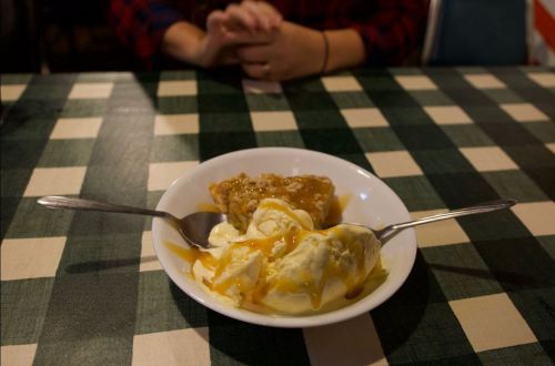 A bowl of vanilla ice cream topped with caramel sauce, served with a slice of dessert on a checkered tablecloth.