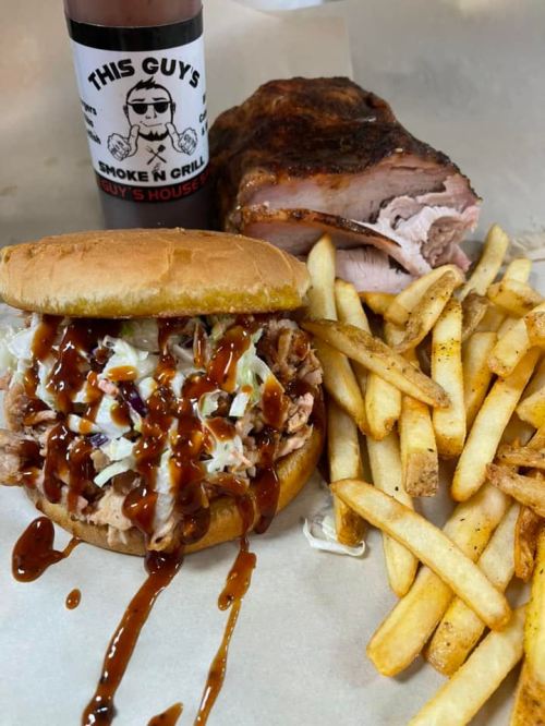 A pulled pork sandwich with BBQ sauce and coleslaw, served with fries and a bottle of BBQ sauce in the background.